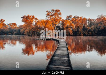 Eine schmale Holzbrücke erstreckt sich über einen ruhigen Teich, der in leuchtenden Herbstfarben reflektiert. Die ruhige Szene fängt die Schönheit der Natur an einem frischen Morgen in Vestec in der Nähe von Prag ein. Stockfoto