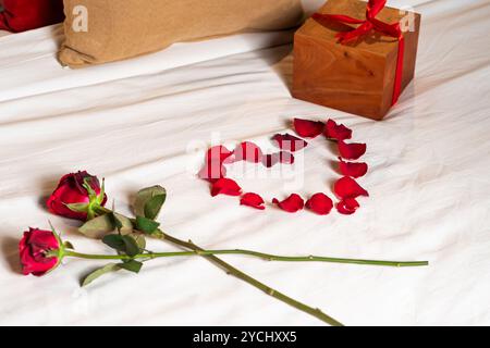 Romantische Einrichtung mit zwei roten Rosen und einer Herzform aus Rosenblättern auf weißem Bettlaken. Eine hölzerne Geschenkbox mit einem roten Band wird nea platziert Stockfoto