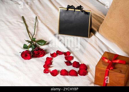 Eine romantische Einrichtung mit zwei roten Rosen, einer Herzform aus Rosenblättern, einer Geschenkbox mit rotem Band und einer schwarzen Geschenktüte auf einer weißen Tagesdecke. Stockfoto