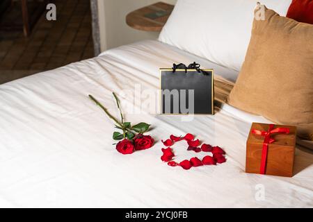 Eine romantische Einrichtung mit zwei roten Rosen, einer Herzform aus Rosenblättern, einer Geschenkbox mit rotem Band und einer schwarzen Geschenktüte auf einer weißen Tagesdecke. Stockfoto