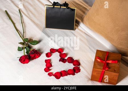 Eine romantische Einrichtung mit zwei roten Rosen, einer Herzform aus Rosenblättern, einer Geschenkbox mit rotem Band und einer schwarzen Geschenktüte auf einer weißen Tagesdecke. Stockfoto