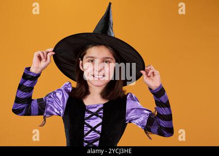 Niedliches lächelndes Mädchen in Hexenkleid und schwarzem Hut, das isoliert vor orangem Hintergrund steht und in die Kamera blickt Stockfoto