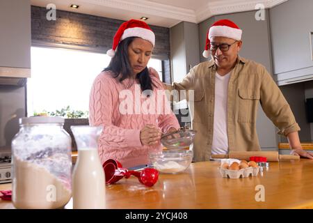 Weihnachten, weihnachtsmütze tragen, multirassisches Seniorenpaar, das in der Küche backen kann, zu Hause Stockfoto