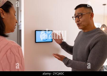 Ich erkläre den intelligenten Thermostat, ein asiatischer Mann, der mit seiner Frau zu Hause über die Temperatureinstellungen spricht Stockfoto