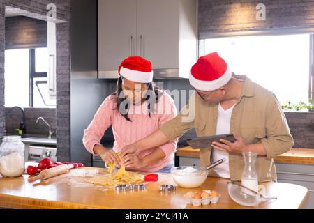 Multirassisches Seniorenpaar, das Weihnachtskekse in der Küche backt, festliche Weihnachtsmützen trägt, zu Hause Stockfoto