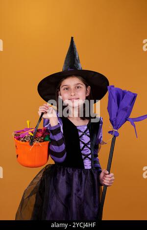 Niedliche kleine Hexe im Halloween-Kostüm, die einen orangen Eimer mit Leckereien und einen lila Besen hält, während sie vor der Kamera steht Stockfoto