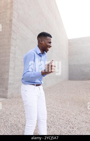 Lachender stilvoller afroamerikanischer Mann mit weißer Jeans und blauem Hemd im Freien, Betonwand auf Hintergrund. Modischer Mittelalter-Mann in seinen 40ern Stockfoto