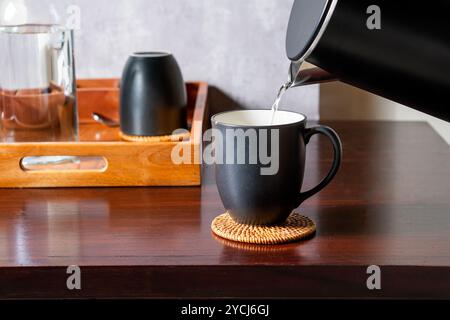 Eine schwarze Kaffeetasse auf einer gewebten Untersetzer, in der heißes Wasser aus einem Wasserkocher gegossen wird. Im Hintergrund befindet sich ein Holztablett mit einem Glas und einem Stockfoto