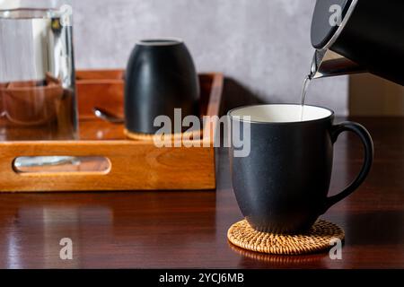 Eine schwarze Kaffeetasse auf einer gewebten Untersetzer, in der heißes Wasser aus einem Wasserkocher gegossen wird. Im Hintergrund befindet sich ein Holztablett mit einem Glas und einem Stockfoto