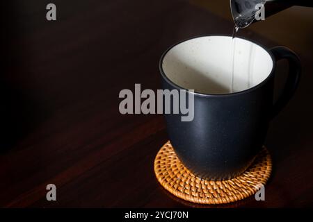 Eine schwarze Kaffeetasse auf einer gewebten Untersetzer, in der heißes Wasser aus einem Wasserkocher gegossen wird. Im Hintergrund befindet sich ein Holztablett mit einem Glas und einem Stockfoto