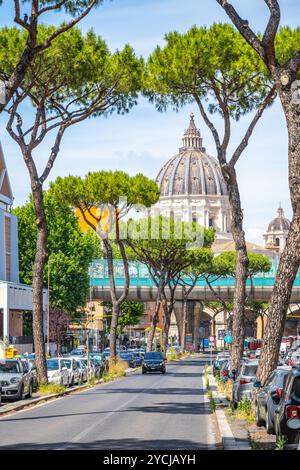 Die majestätische Kuppel des Petersdoms erhebt sich über der von Bäumen gesäumten Straße und zeigt die lebhafte Atmosphäre der Vatikanstadt an einem sonnigen Tag zwischen Autos und blauem Himmel. Stockfoto