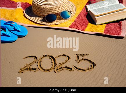 2025 Text Handschrift auf dem Sand und ein Strandtuch mit Hut, Sonnenbrille und Flip Flop - Konzept Neujahr Stockfoto