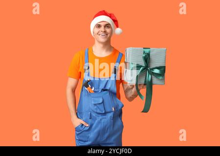Männlicher Klempner im Weihnachtsmann-Hut mit Werkzeugen und Geschenkbox auf orangem Hintergrund Stockfoto