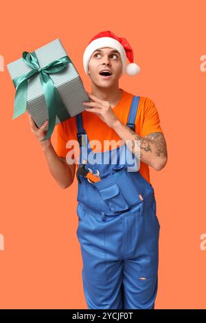 Männlicher Klempner im Weihnachtsmann-Hut mit Werkzeugen und Geschenkbox auf orangem Hintergrund Stockfoto
