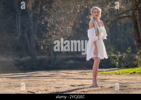 Eine atemberaubende junge Frau geht an einem Bach vorbei, ihr weißes Frühlingskleid schwingt anmutig im Wind. Umgeben von üppigen Wäldern, genießt sie die Ruhe Stockfoto