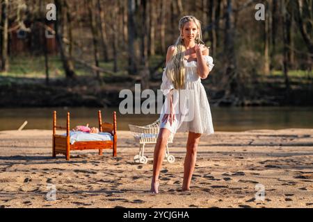 Eine atemberaubende junge Frau geht an einem Bach vorbei, ihr weißes Frühlingskleid schwingt anmutig im Wind. Umgeben von üppigen Wäldern, genießt sie die Ruhe Stockfoto