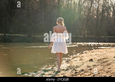 Eine atemberaubende junge Frau geht an einem Bach vorbei, ihr weißes Frühlingskleid schwingt anmutig im Wind. Umgeben von üppigen Wäldern, genießt sie die Ruhe Stockfoto