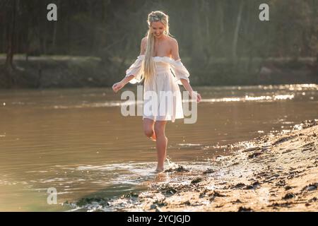 Eine atemberaubende junge Frau geht an einem Bach vorbei, ihr weißes Frühlingskleid schwingt anmutig im Wind. Umgeben von üppigen Wäldern, genießt sie die Ruhe Stockfoto