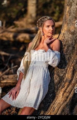 Eine atemberaubende junge Frau geht an einem Bach vorbei, ihr weißes Frühlingskleid schwingt anmutig im Wind. Umgeben von üppigen Wäldern, genießt sie die Ruhe Stockfoto