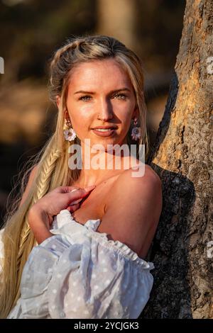 Eine atemberaubende junge Frau geht an einem Bach vorbei, ihr weißes Frühlingskleid schwingt anmutig im Wind. Umgeben von üppigen Wäldern, genießt sie die Ruhe Stockfoto