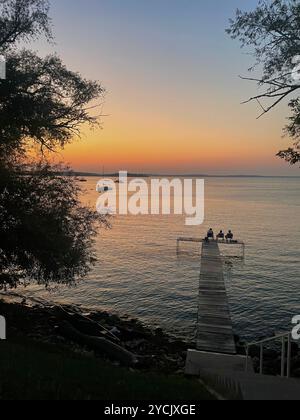 Eine ruhige Seeszene bei Sonnenuntergang fängt eine Gruppe von Menschen ein, die zusammen auf einem hölzernen Dock sitzen Stockfoto