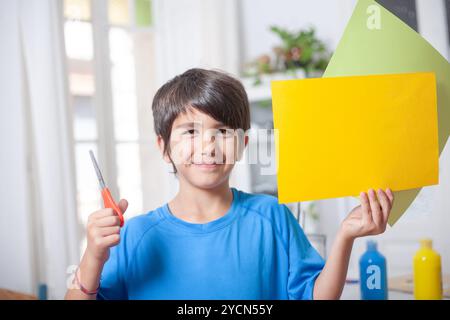 Ein kleiner Junge hält ein gelbes Stück Papier und eine Schere. Er lächelt und freut sich, die Zeitung zu schneiden. Die Szene deutet darauf hin Stockfoto