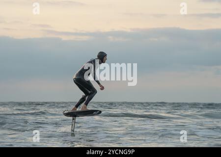 Ein junger Mann in einem schwarzen Neoprenanzug reitet auf einem Tragflächenboot-Surfbrett in kleinen Wellen vor dem Strand von Bourenmouth. Stockfoto