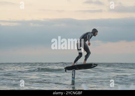 Ein junger Mann in einem schwarzen Neoprenanzug reitet auf einem Tragflächenboot-Surfbrett in kleinen Wellen vor dem Strand von Bourenmouth. Stockfoto