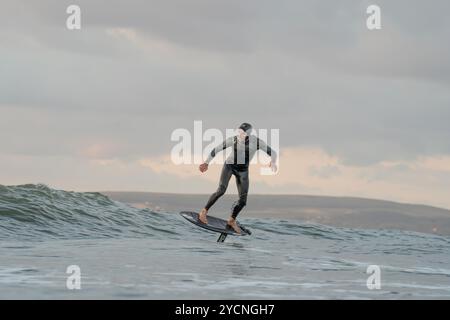 Ein junger Mann in einem schwarzen Neoprenanzug reitet auf einem Tragflächenboot-Surfbrett in kleinen Wellen vor dem Strand von Bourenmouth. Stockfoto