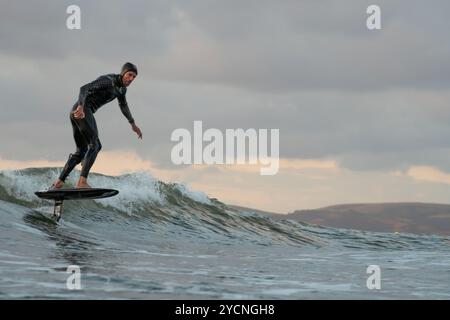 Ein junger Mann in einem schwarzen Neoprenanzug reitet auf einem Tragflächenboot-Surfbrett in kleinen Wellen vor dem Strand von Bourenmouth. Stockfoto