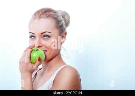 Fröhliche junge Frau mit blondem Haar in einem Brötchen isst glücklich einen grünen Apfel, strahlt Glück aus und genießt einen gesunden Snack Stockfoto