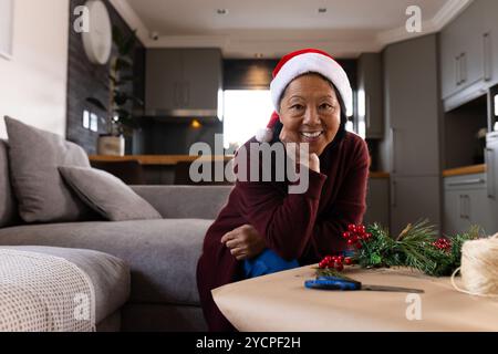 Lächelnde asiatische Seniorin mit Weihnachtsmütze, die Weihnachtsdekorationen zuhause vorbereitet Stockfoto