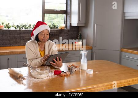 Asiatische Seniorin im Weihnachtsmann-Hut, die Tablette benutzt, während sie zu Hause Weihnachtskekse backen Stockfoto