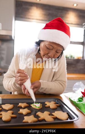 Mit Weihnachtsmütze, asiatische Seniorin, die Weihnachtskekse zu Hause dekoriert, konzentriert Stockfoto