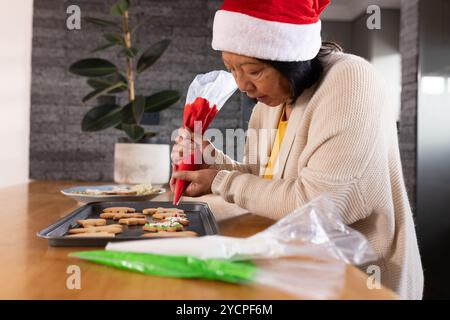 asiatische Seniorin, die Weihnachtskekse zu Hause dekoriert, mit Weihnachtsmütze Stockfoto