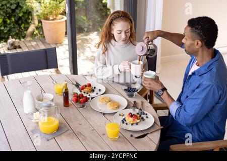 Ein multirassisches Paar genießt zu Hause ein gemeinsames Frühstück, gießt Kaffee und lächelt Stockfoto