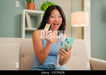 Begeisterte junge asiatische Frau, die zu Hause mit Smartphone und Kreditkarte online einkaufen kann. Glückliche Kunden, die Lieferung bestellen oder online bezahlen, genießen bequemen E-Commerce. Hochwertige Fotos Stockfoto