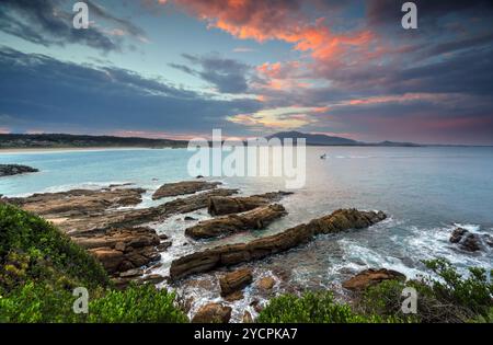 Finger aus Felsen erstrecken sich bei Sonnenuntergang vom Dickinson Park in den Ozean. Der Berg Gulaga erhebt sich in der Ferne und ein Boot kehrt zum Hafen zurück. Stockfoto