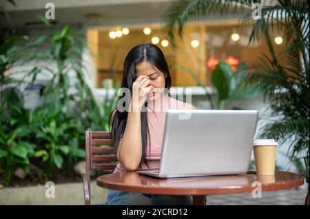 Eine ernsthafte, nachdenkliche asiatische Frau arbeitet aus der Ferne von einem Café aus, sitzt mit ihrem Laptop an einem Tisch im Freien und fühlt sich müde und besorgt über abo Stockfoto