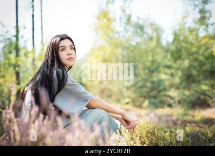 Eine junge Frau sitzt ruhig auf dem Waldboden, ihr langes Haar zieht sich über den Rücken. Sie erscheint tief in Gedanken, umgeben von blühenden Wildblumen Stockfoto