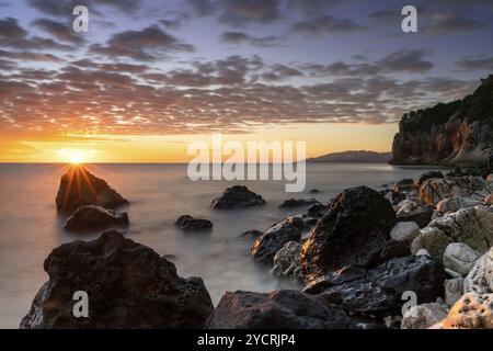 Ein farbenfroher Sonnenaufgang in Cala Gonone mit schwarz-weißen Felsen und Felsbrocken und einem Sonnenaufgang Stockfoto