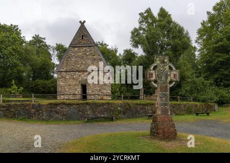 Wexford, Irland, 18. August 2022: Blick auf ein früh rekonstruiertes christliches Kloster im Irish National Heritage Park mit einem großen keltischen Kreuz Stockfoto