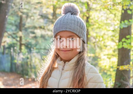 Ein 6-jähriges Mädchen mit grauem Hut und Jacke lächelt in einem sonnendurchfluteten Herbstwald. Stockfoto