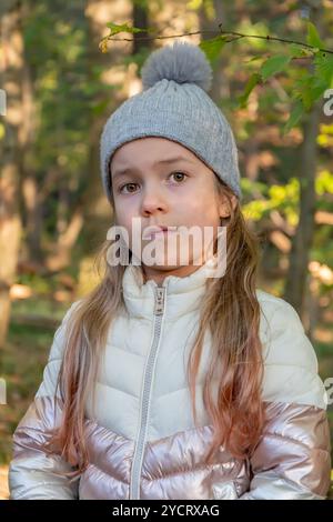 Ein kleines Mädchen von 6-8 Jahren mit grauem Hut und weißer Jacke steht nachdenklich zwischen den Bäumen in einem ruhigen Wald an einem sonnigen Herbsttag. Stockfoto