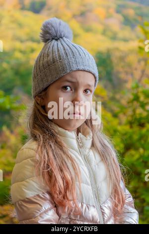 Ein kleines Mädchen von 6-8 Jahren mit grauem Hut und weißer Jacke steht nachdenklich zwischen den Bäumen in einem ruhigen Wald an einem sonnigen Herbsttag. Stockfoto