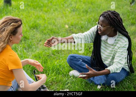 Zwei junge Frauen, wahrscheinlich Studenten, sitzen auf Gras in einem Park und führen eine Diskussion. Eine Frau macht Gesten mit einem Zweig, als sie ihrem Freund etwas erklärt Stockfoto