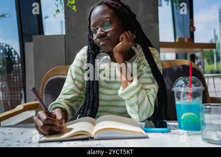Die junge schwarze Frau macht sich gedankenvoll Notizen in ihr Notizbuch, während sie an einem Tisch in einem Café sitzt. Sie trägt ein grün-weiß gestreiftes Hemd und hat Kopfhörer um den Hals Stockfoto