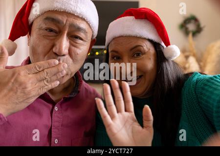 Chrsitmas Zeit, auf Videoanruf, multirassisches Seniorenpaar mit weihnachtsmützen, die zu Hause Küsse blasen Stockfoto