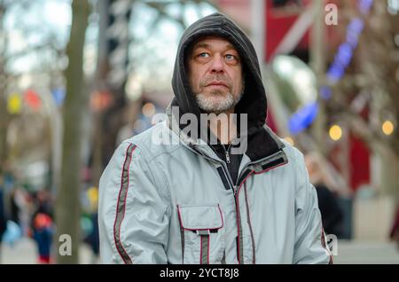 Straßenporträt eines älteren Mannes von 50-55 Jahren, der ein Kapuzenpullover auf dem Kopf auf einem verschwommenen urbanen Hintergrund trägt. Stockfoto