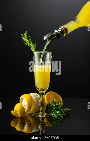 Traditioneller italienischer Likör Limoncello mit Rosmarinzweig auf schwarzem reflektierendem Hintergrund. Alkohol wird aus einer Flasche in ein Glas gegossen. Stockfoto